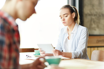 Fototapeta premium Casual teen girl watching tablet sitting at table with coffee and anonymous students