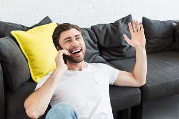 Fototapeta premium young man gesturing by hand and talking on smartphone near couch at home