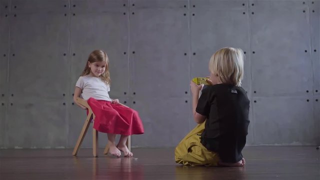 Blond little boy sitting on the floor is taking a picture of a little girl sitting on a chair in a gray wall room. Concept of art and prodigy creative photographer. Locked down slow motion medium shot
