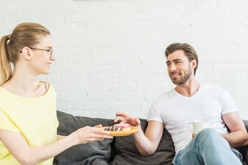 smiling woman in eyeglasses offering donuts to boyfriend with coffee cup at home