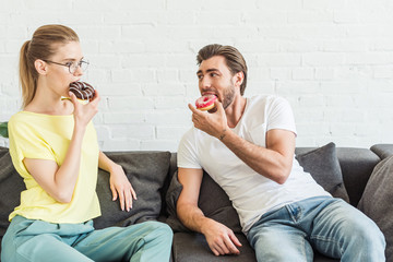 young couple eating donuts and sitting on sofa at home