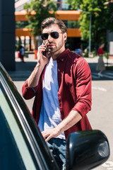 stylish man in sunglasses talking on smartphone and opening door of own car at urban street