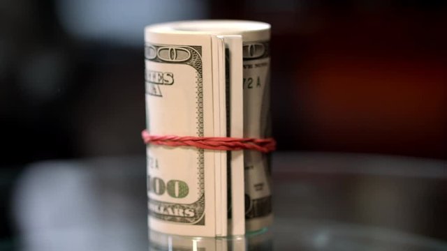 Rotating roll of hundred USA dollar bills with red rubber band. Cash money roll on table. Family savings concept. Starting investment capital. Exchange currency financial operations
