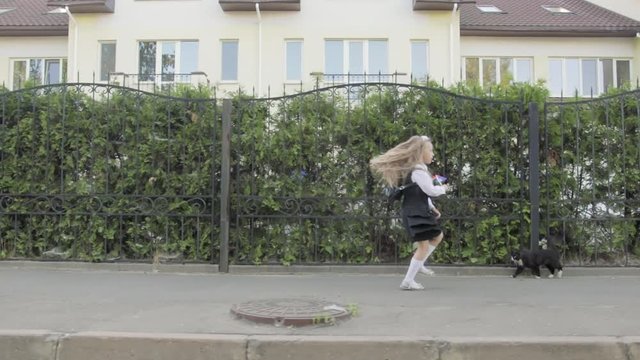 Little funny schoolgirl in school uniform and briefcase on his shoulders walking around the area. In the hands of the girl rainbow spinner, near the road is a cat. Steadicam shot