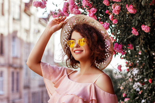 Outdoor close up portrait of young beautiful happy smiling curly girl wearing stylish yellow sunglasses, straw hat, pink top with ruffles. Model posing near blooming roses. Summer fashion concept
