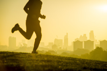 Defocused silhouette of jogger running on a green grassy hill in front of the misty London city skyline. Focus on buildings in the background.