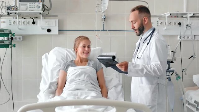 Male doctor talking to female patient in hospital bed. Smiling doctor with clipboard and x-ray picture attending sick woman in hospital ward