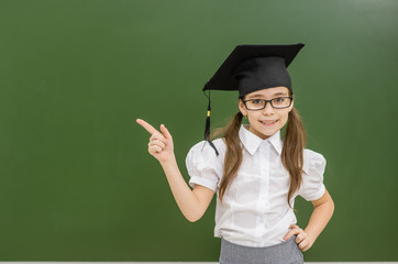 Happy girl in graduation cap standing near a school board and pointing away on empty space. Space...