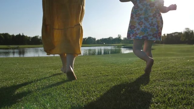 Close-up legs of mother and her little daughter running barefoot on green grass in summer during sunset. Joyful family running to pond together and having fun during holiday. Slow motion steadicam