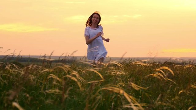 Beautiful girl wearing white dress running through beautiful field at sunset. Young woman jogging at the meadow and enjoying freedom. Summer leisure at nature concept.