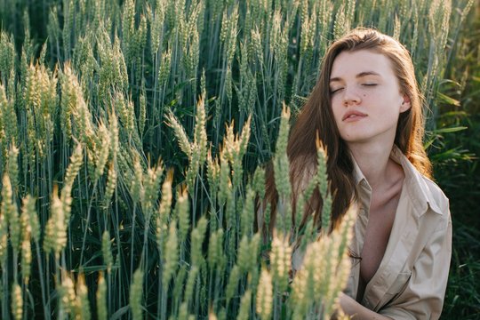 Portrait of young attractive female daydreaming in the field of green wheat with closed eyes