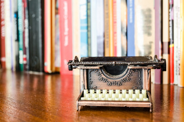 Small typewriter in copper tones in focus ahead of books. Decoration item. Concept of reading, writing, old times, past.