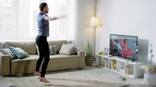 Wide shot of laughing bearded man in sportswear doing scissor jumps and jumping jacks while looking at cheerful online person coach on TV screen