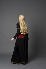 Fototapeta premium full length portrait of pretty blonde lady wearing a red and black fantasy medieval gown. standing pose on grey background.