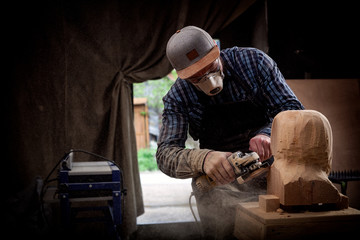 Experienced carpenter in work clothes and small buiness owner working in woodwork workshop, processes the board with an angle grinder on the table is a hammer and many tools