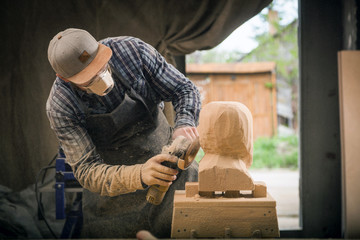 A young man carpenter in a suit, goggles   saws a man's head with a tree , using an angle grinder  in the workshop, around a lot of tools for work
