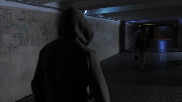 Woman walking alone in mini skirt in dark underpass and male figure in hoodie starts chasing her at night. Criminal following victim in dark subway with intention of murder or abuse. Steadicam shot