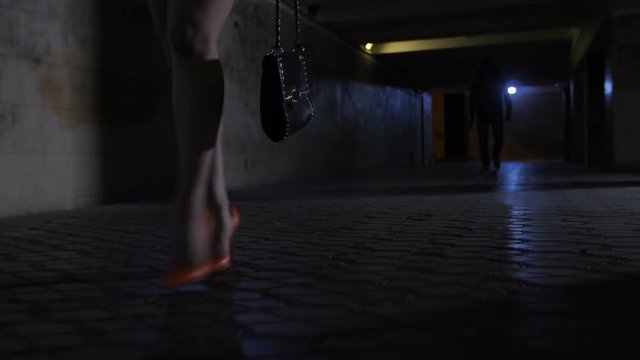 Female slim legs in high heels and mini skirt walking on the foreground in dark underpass subway at night. Male silhouette walking on the background chasing and following lonely female