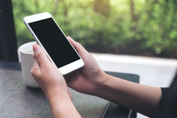 Mockup image of hands holding white mobile phone with blank black desktop screen with coffee cup on table in cafe