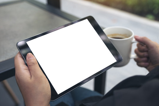 Mockup image of a woman sitting cross legged and holding black tablet pc with blank white desktop screen while drinking coffee in cafe