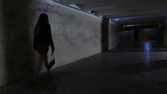 Drunk boozy female in mini skirt and high heeled shoes walking alone in dark underpass tunnel alone. Tipsy woman stumbling and touching the wall trying to find the way home at night