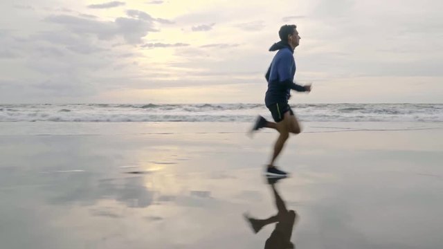Side view of sporty man in hoodie and shorts running on sand and listening to music when warming up before summer workout at the beach