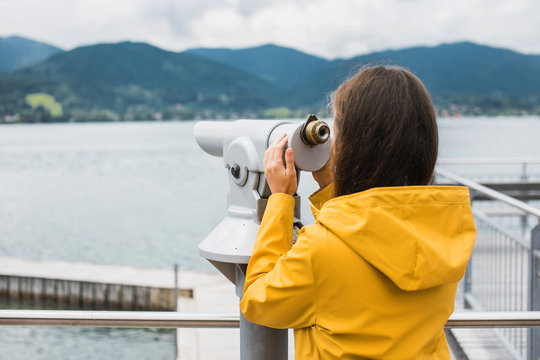 Girl in a yellow raincoat looking through coin-operated telescope at the opposite bank of the lake in Alps