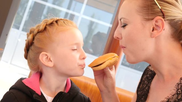 Little kid girl with her mother funny bite and eat a burger in street fast food restaurant