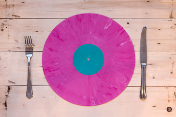 Pink vinyl record in the form of a plate with fork and knife on a wooden table background. Music...