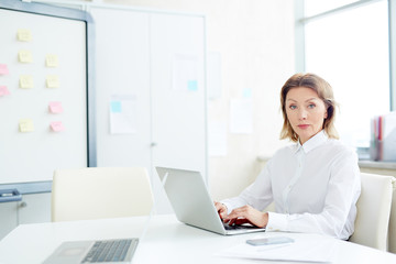Portrait of confident mature businesswoman looking at camera while typing on laptop at office desk