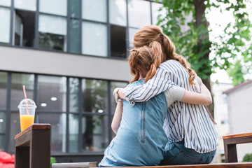back view of mother and daughter hugging each other while resting on bench on street