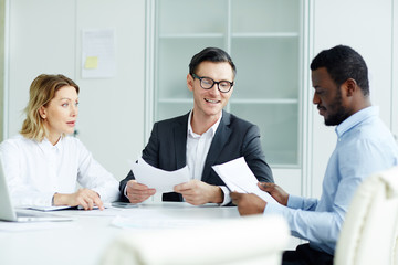 Adult woman and two multiethnic men in classical clothes sitting at white table and discussing with papers in hands in working space