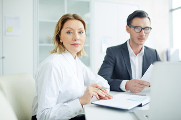 Businesswoman in elegant clothes and notebook in office with man behind and discussing contracts