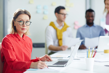 Serious businesswoman using laptop and writing at table in office with people on background