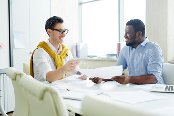 Fototapeta premium Cheerful stylish multiracial men chatting at table in light office and discussing papers while working. 