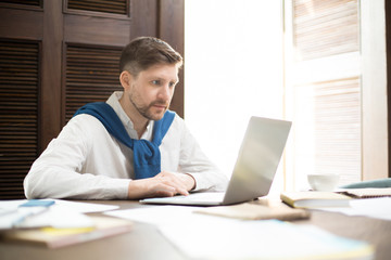 Fototapeta premium Attractive businessman sitting at cafe table amidst various papers and browsing laptop. 