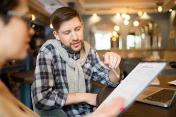 Handsome man in casual clothes sitting at cafe table and pointing at important items in clipboard document for colleague. 