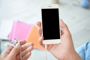 Young woman holding mobile phone with blank screen and earphones in hands, indoors