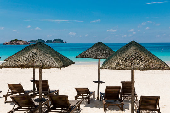 Typical holiday beach scenery with wooden sun loungers under the shade of wooden umbrellas with thatched roofs in the middle of powdery white sand and turquoise blue water on Redang Island, Malaysia. 