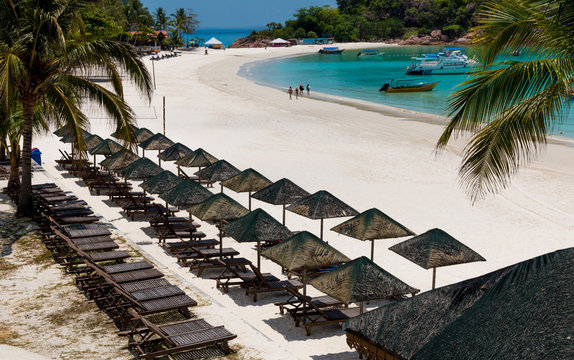 Lovely holiday beach scenery with rows of wooden sun loungers and umbrellas with thatched roofs. Tourists walking along powdery white sand and boats anchored on turquoise blue water of Redang Island.