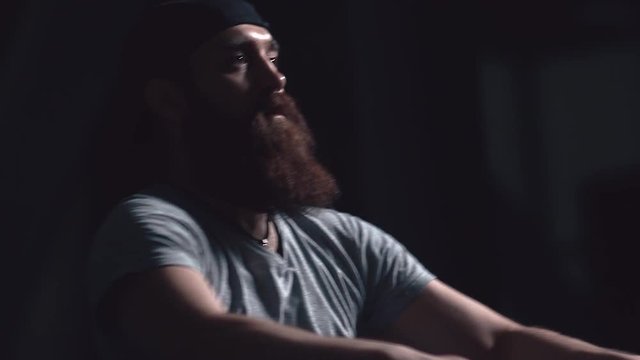 Portrait of an athlete: a brutal man with a beard is engaged in a rowing machine, he makes a powerful jerk. A man's face while doing sports. Shooted against a dark background. Sport in slowmotion.