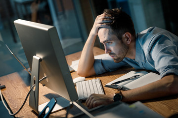 Young man leaning on hand looking extremely tired while finishing overtime project watching...
