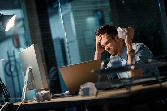 Angry man unhappy with failures sitting at table and crumpling papers in office watching laptop