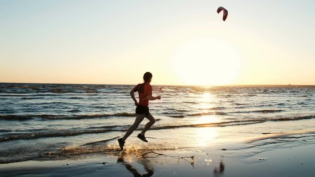 Man runs along the beach and is happy at the finish on the background of kitesurfers at sunset. Athlete splashes water