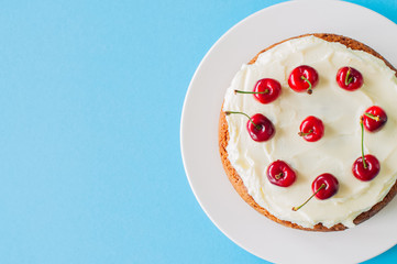 Homemade sour cream cake with cream cheese frosting decorated with fresh ripe cherries on a light blue background.