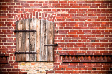 Closed brown red wooden window with rusted metal hinges on a grungy red brick wall of an old barn weathered by exposure to the elements.