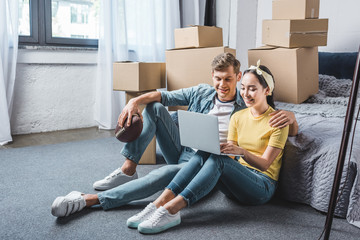 happy young couple sitting on floor of bedroom and using laptop while moving into new home