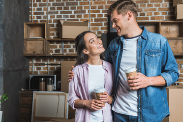 young interracial couple with paper cups of coffee standing on kitchen while moving into new home and looking at each other