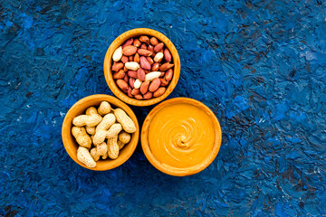 Peanut paste concept. Bowls with butter, nuts in shell, peeled nuts on blue background top view copy space