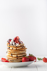 Stack of pancakes with strawberries, whip cream and chocolate syrup on a white plate on a white background. Copy space.